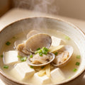 Clams and tofu simmering in a light broth in a ceramic bowl